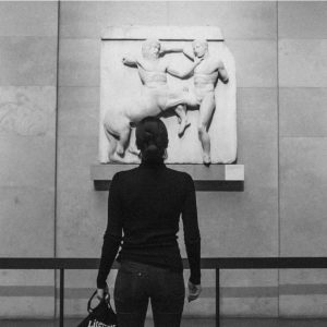 A woman stands in front of a sculpture, depicting a centaur strangling a man.